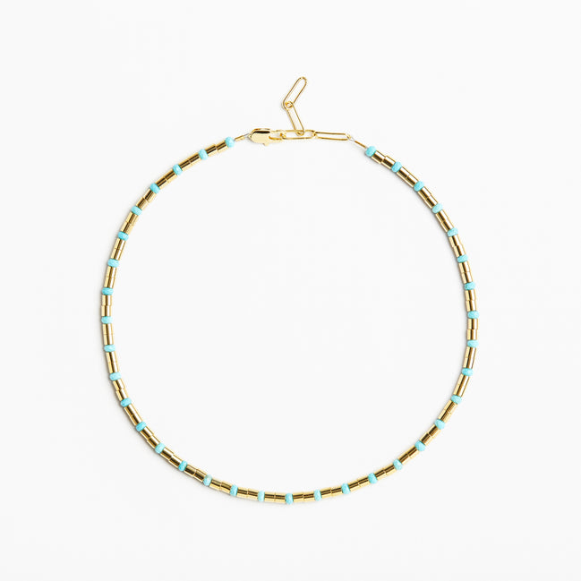 A delicate gold chain necklace with small, evenly spaced light blue beads, displayed in a circular shape against a clean white background.