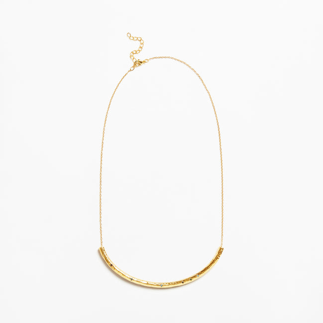 A delicate gold chain necklace featuring a textured, curved bar pendant at the bottom, displayed against a clean white background.