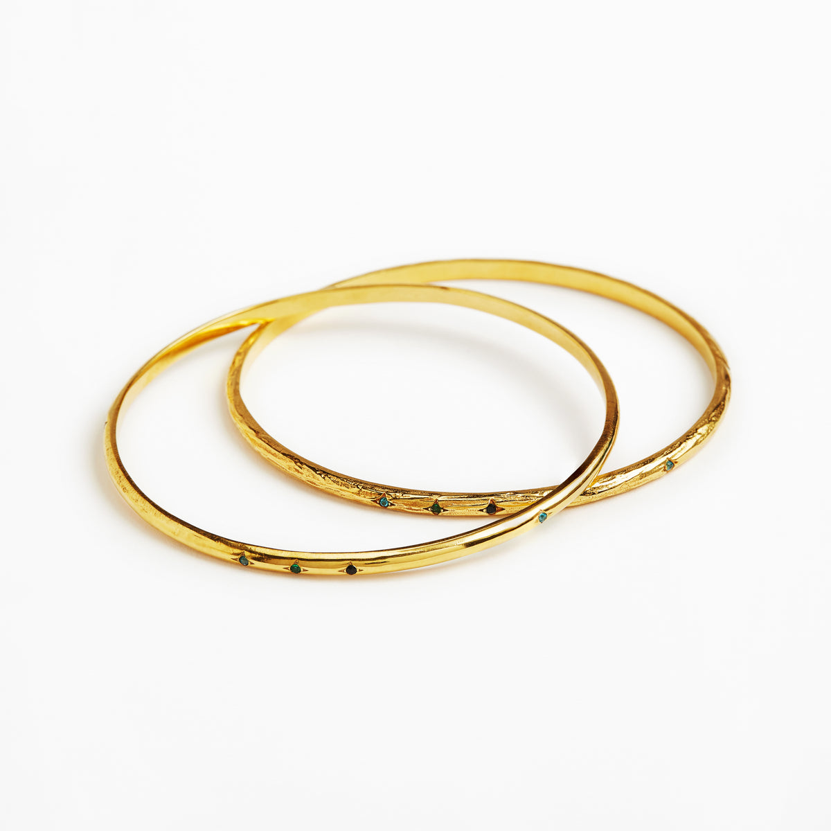 Two thin gold bangles with small dark green stone accents, overlapping on a plain white background.