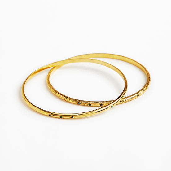 Two thin gold bangles with small dark green stone accents, overlapping on a plain white background.