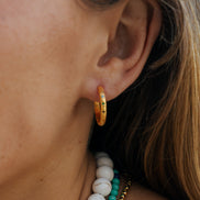 A close-up of a person's ear wearing a small gold hoop earring with colorful enamel dots, along with layered white and turquoise beaded necklaces.