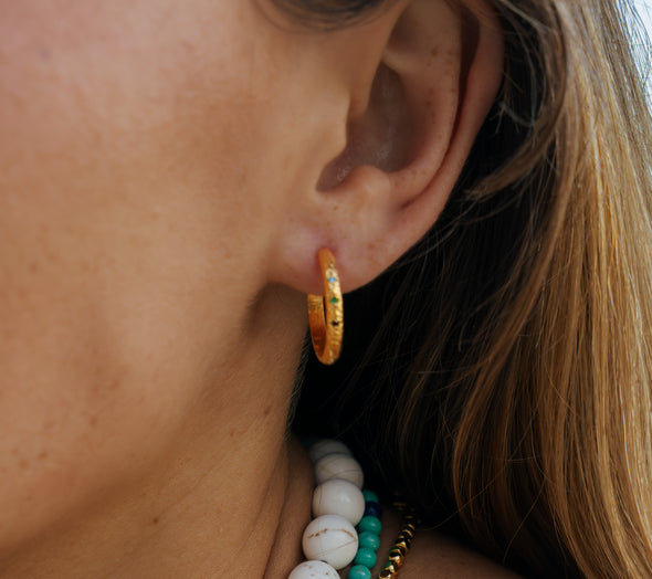 A close-up of a person's ear wearing a small gold hoop earring with colorful enamel dots, along with layered white and turquoise beaded necklaces.