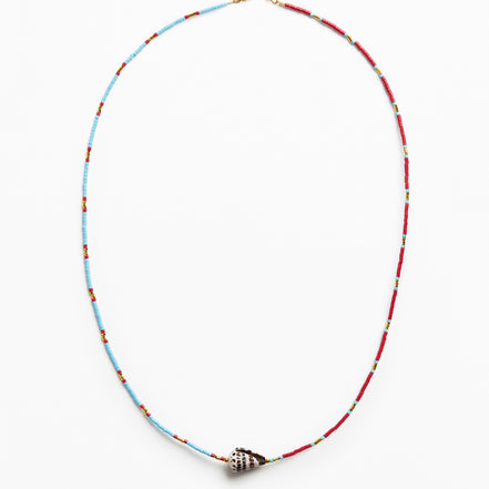 A delicate beaded necklace with alternating light blue and red beads, featuring a small metallic shell-shaped charm at the center, displayed on a clean white background.