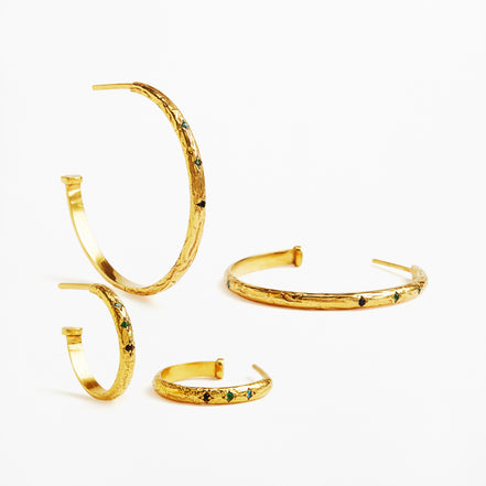 Four textured gold hoop and cuff earrings with small green and black stone accents, arranged on a clean white background.