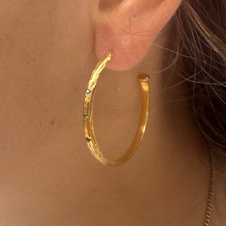 A close-up of a textured gold hoop earring with small green and blue gemstones, worn on a person's ear.
