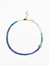 A beaded necklace with blue, teal, and white beads, accented by a small silver-toned skull charm and gold-toned clasp, shown on a plain white background.