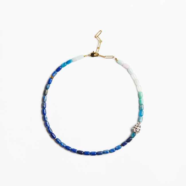 A beaded necklace with blue, teal, and white beads, accented by a small silver-toned skull charm and gold-toned clasp, shown on a plain white background.