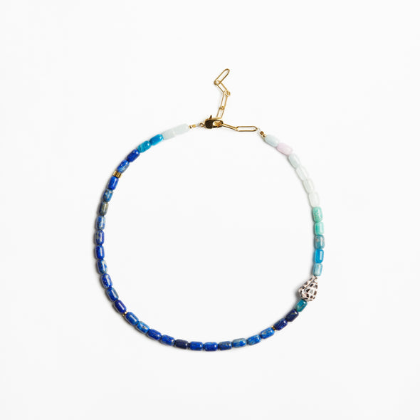 A beaded necklace with blue, teal, and white beads, accented by a small silver-toned skull charm and gold-toned clasp, shown on a plain white background.