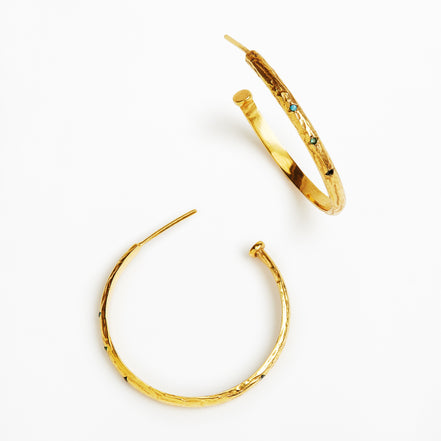 A pair of textured gold hoop earrings with small dark and light blue gemstone accents, displayed on a clean white background.