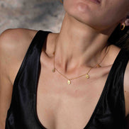 A close-up lifestyle shot of a model wearing a delicate gold necklace with small disc charms, paired with a black satin top against a sandy beach background.