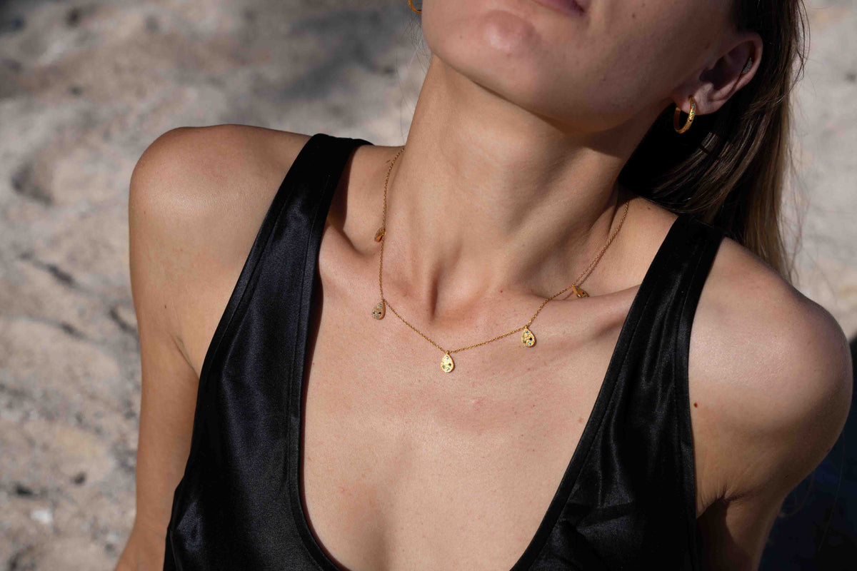 A close-up lifestyle shot of a model wearing a delicate gold necklace with small disc charms, paired with a black satin top against a sandy beach background.