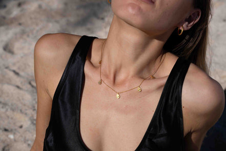 A close-up lifestyle shot of a model wearing a delicate gold necklace with small disc charms, paired with a black satin top against a sandy beach background.