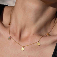 A close-up lifestyle shot of a model wearing a delicate gold necklace with small disc charms, paired with a black satin top against a sandy beach background.