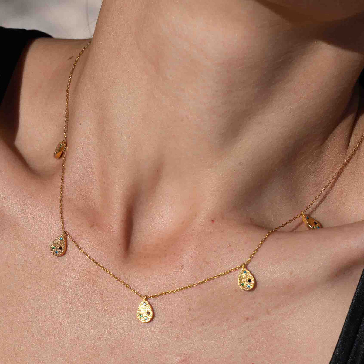 A close-up lifestyle shot of a model wearing a delicate gold necklace with small disc charms, paired with a black satin top against a sandy beach background.