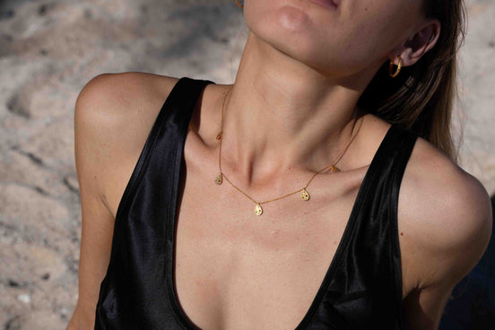 A close-up lifestyle shot of a model wearing a delicate gold necklace with small disc charms, paired with a black satin top against a sandy beach background.