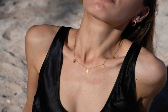 A close-up lifestyle shot of a model wearing a delicate gold necklace with small disc charms, paired with a black satin top against a sandy beach background.