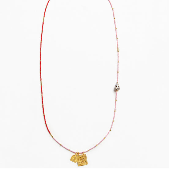 A delicate beaded necklace featuring a red and light pink string with small metallic beads, a silver-toned bell-shaped charm, and two gold-toned pendant tags at the bottom, displayed on a clean white background.