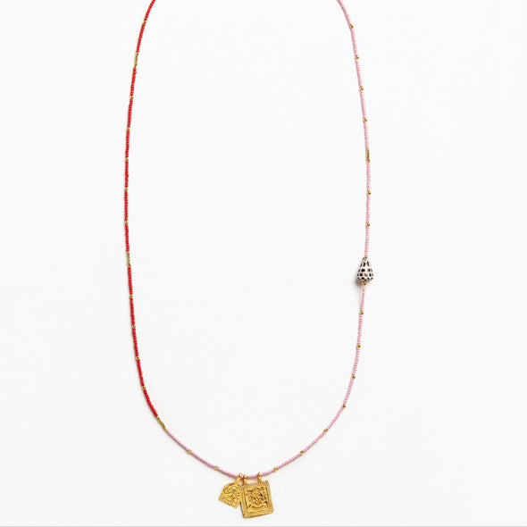 A delicate beaded necklace featuring a red and light pink string with small metallic beads, a silver-toned bell-shaped charm, and two gold-toned pendant tags at the bottom, displayed on a clean white background.