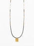 A long beaded necklace featuring a black cord, white and black beads, and a small gold square pendant with an engraved design, shown against a plain white background.