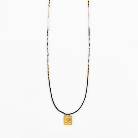 A long beaded necklace featuring a black cord, white and black beads, and a small gold square pendant with an engraved design, shown against a plain white background.