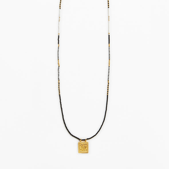 A long beaded necklace featuring a black cord, white and black beads, and a small gold square pendant with an engraved design, shown against a plain white background.