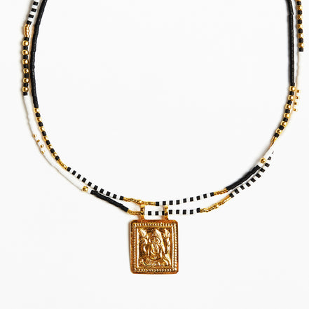 A beaded necklace featuring black and white beads with gold-toned chain accents and a square gold pendant with an embossed figure, shown against a plain white background.