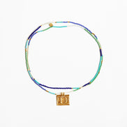 A beaded necklace with multiple strands of blue, green, and white beads, featuring a gold square pendant with an embossed figure, displayed on a clean white background.