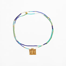 A beaded necklace with multiple strands of blue, green, and white beads, featuring a gold square pendant with an embossed figure, displayed on a clean white background.