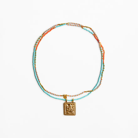 A delicate beaded necklace with turquoise, orange, and gold beads, featuring a square gold pendant with an embossed design, displayed on a clean white background.