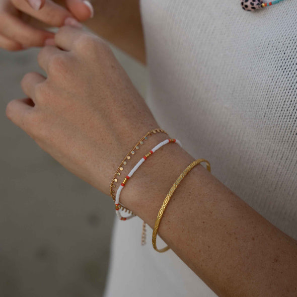 A close-up of a person's wrist wearing three delicate bracelets: a textured gold bangle, a white beaded bracelet with small red accents, and a thin gold chain bracelet.