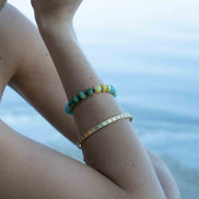 A close-up of a person's wrist wearing two bracelets: a turquoise and yellow beaded bracelet and a gold bangle, with a blurred blue water background.