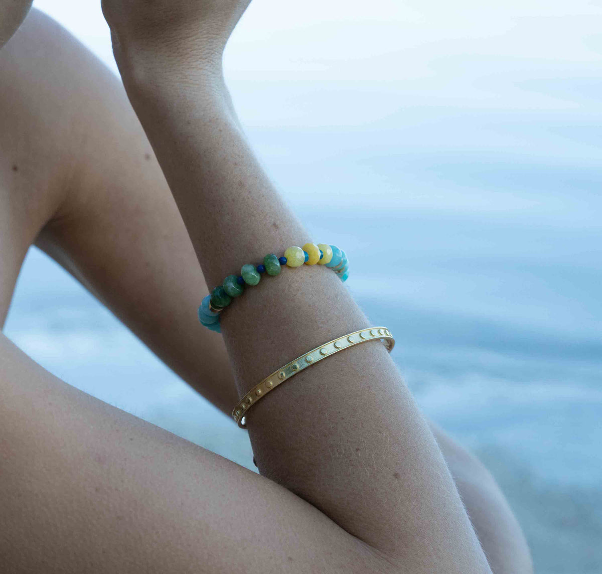 A close-up of a person's wrist wearing two bracelets: a turquoise and yellow beaded bracelet and a gold bangle, with a blurred blue water background.