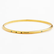 A thin yellow gold bangle bracelet with five small, evenly spaced colored gemstone accents in blue, green, and black, set against a plain white background.