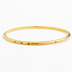 A thin yellow gold bangle bracelet with five small, evenly spaced colored gemstone accents in blue, green, and black, set against a plain white background.