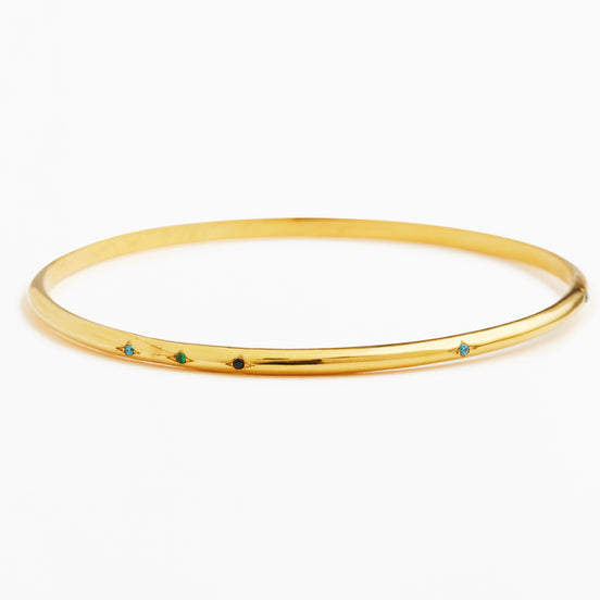 A thin yellow gold bangle bracelet with five small, evenly spaced colored gemstone accents in blue, green, and black, set against a plain white background.
