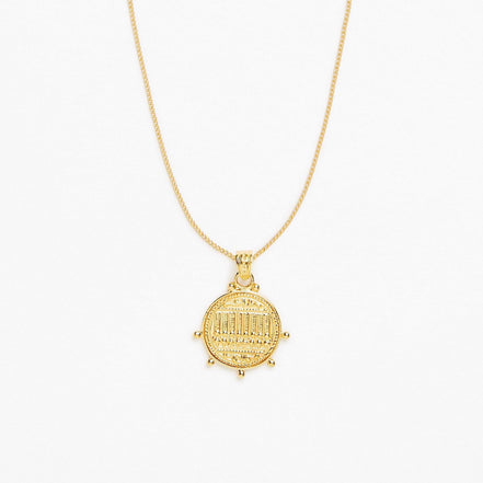 A gold pendant necklace featuring a circular coin-style charm with an engraved arch design, suspended from a delicate gold chain against a plain white background.