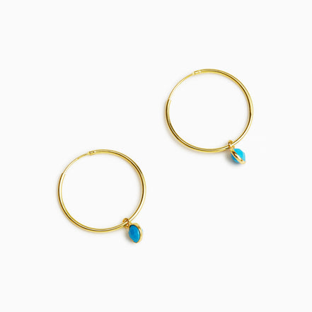 A pair of gold hoop earrings, each adorned with a small teardrop-shaped turquoise stone, displayed on a clean white background.