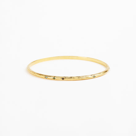 A thin gold bangle bracelet with a textured, hammered finish and small dark green stone accents spaced along the front edge, shown on a plain white background.