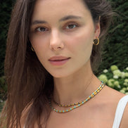 A close-up portrait of a woman wearing a double-strand beaded necklace with yellow, blue, and white beads, along with gold hoop earrings, set against a blurred green garden background.
