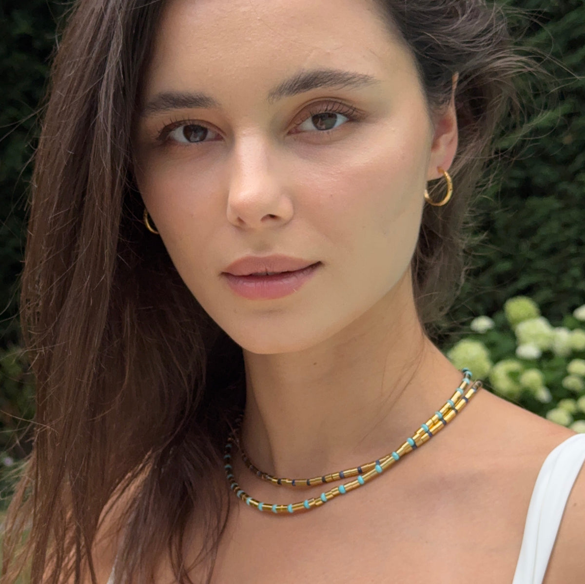 A close-up portrait of a woman wearing a double-strand beaded necklace with yellow, blue, and white beads, along with gold hoop earrings, set against a blurred green garden background.