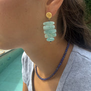A close-up lifestyle shot of a person wearing a gold and light blue stone drop earring, along with a matching blue beaded necklace.