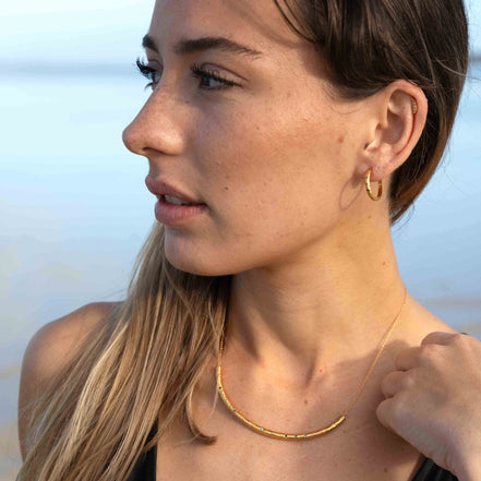 A woman in profile wearing a delicate gold necklace and a small gold hoop earring, photographed outdoors against a soft blurred blue background.