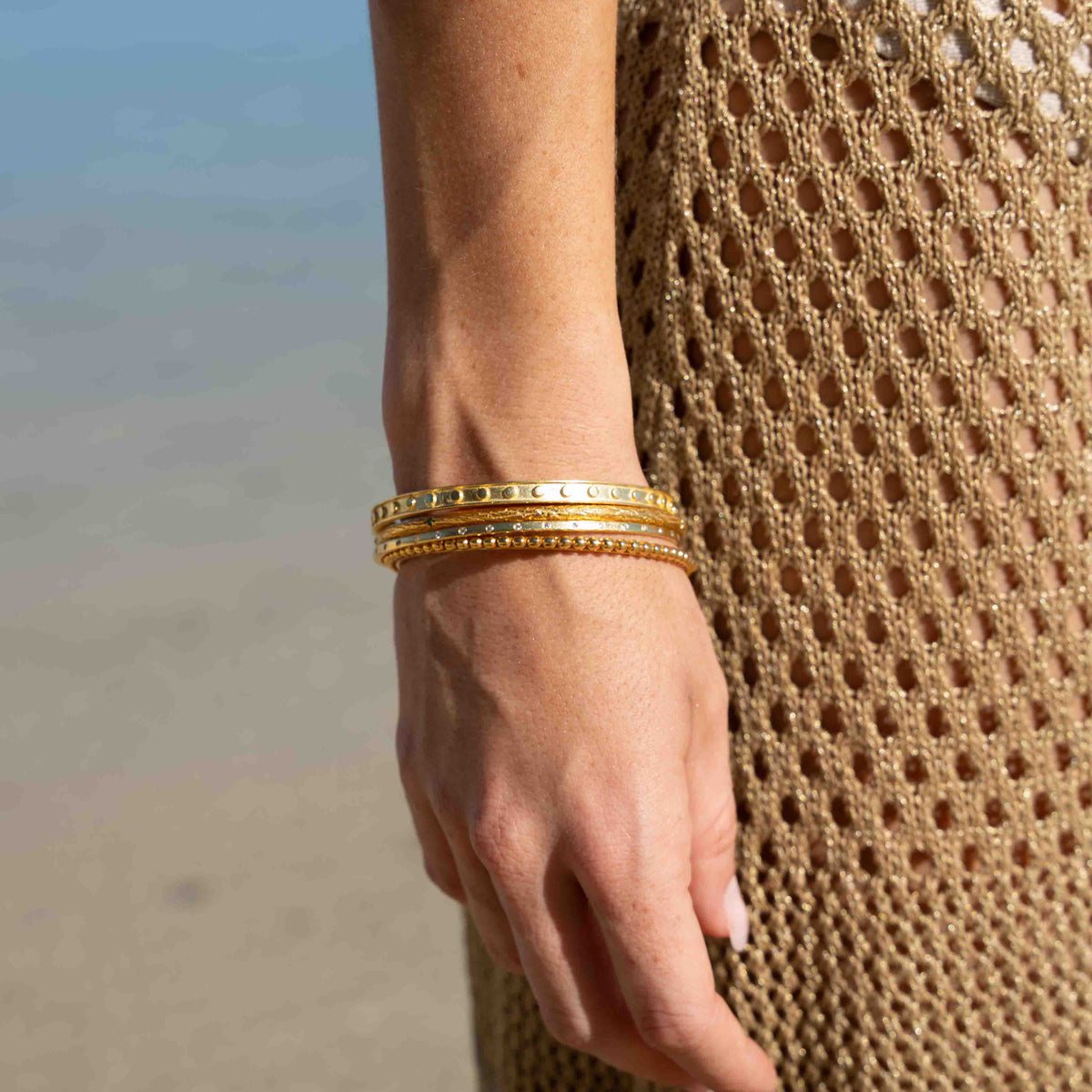 A close-up of a person's wrist wearing three stacked gold bangles, with a sandy beach and ocean in the background and a beige woven dress visible.