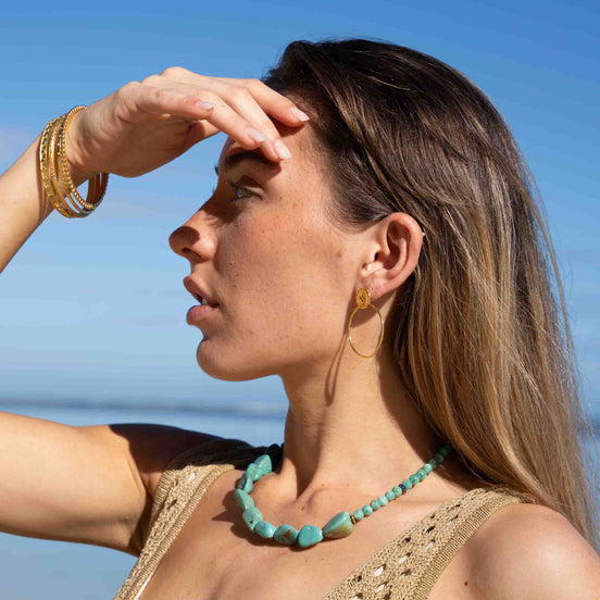 A woman outdoors by the sea wearing a turquoise beaded necklace and gold hoop earrings, with her hand shielding her eyes from the sun against a blue sky.