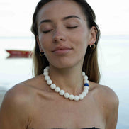 A woman with her eyes closed wears a white beaded necklace with a small blue accent bead, photographed outdoors against a soft, blurred water background.