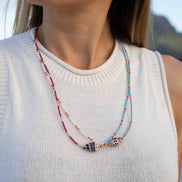 A close-up of a woman wearing a double-strand beaded necklace with shell charms. The necklace features red, light blue, and white beads with small orange accents, and two shell pendants.