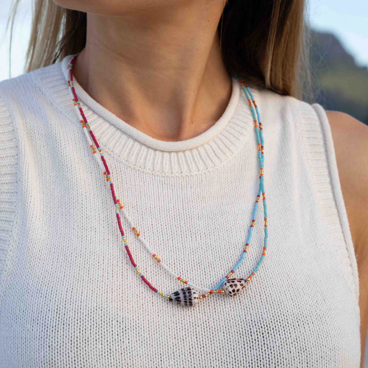 A close-up of a woman wearing a double-strand beaded necklace with shell charms. The necklace features red, light blue, and white beads with small orange accents, and two shell pendants.