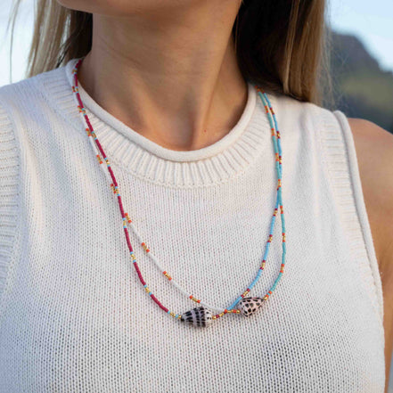 A close-up of a woman wearing a double-strand beaded necklace with shell charms. The necklace features red, light blue, and white beads with small orange accents, and two shell pendants.