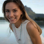 A smiling woman with long light brown hair wearing a white sleeveless knit top and a colorful beaded necklace, photographed outdoors against a soft-focus water and mountain background.