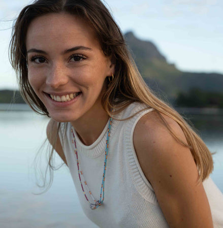 A smiling woman with long light brown hair wearing a white sleeveless knit top and a colorful beaded necklace, photographed outdoors against a soft-focus water and mountain background.
